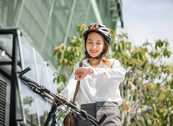 Eine junge Frau mit weißer Bluse und schwarzem Fahrradhelm blickt auf ihre Uhr und fährt mit dem Fahrrad zur Arbeit